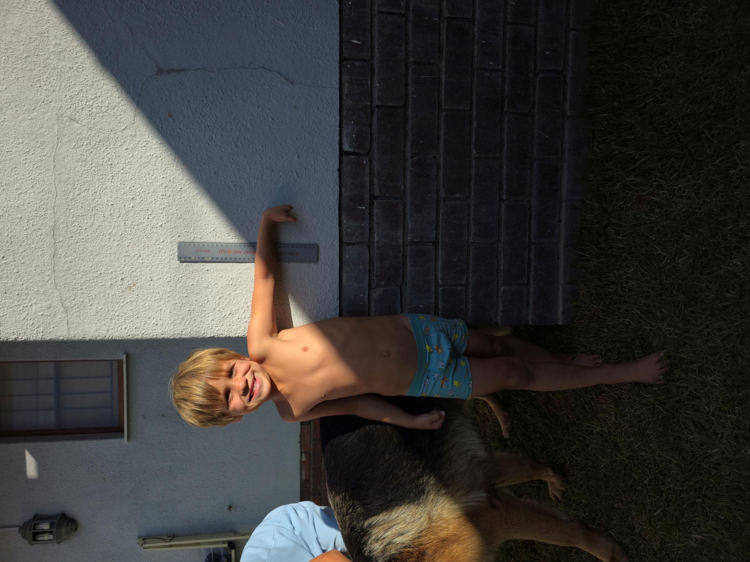 Noah shirtless outside against a white wall with a ruler for scale, left arm extended, measurement photo